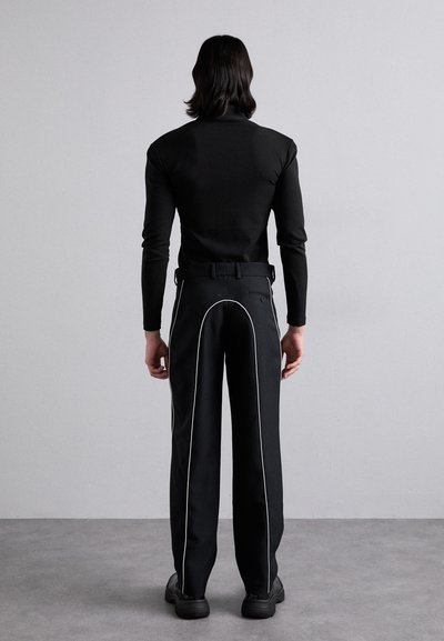 Haut ajusté noir à manches longues, associé à un pantalon noir avec des détails en passepoil blanc sur les côtés, et une ceinture. Fond uni.