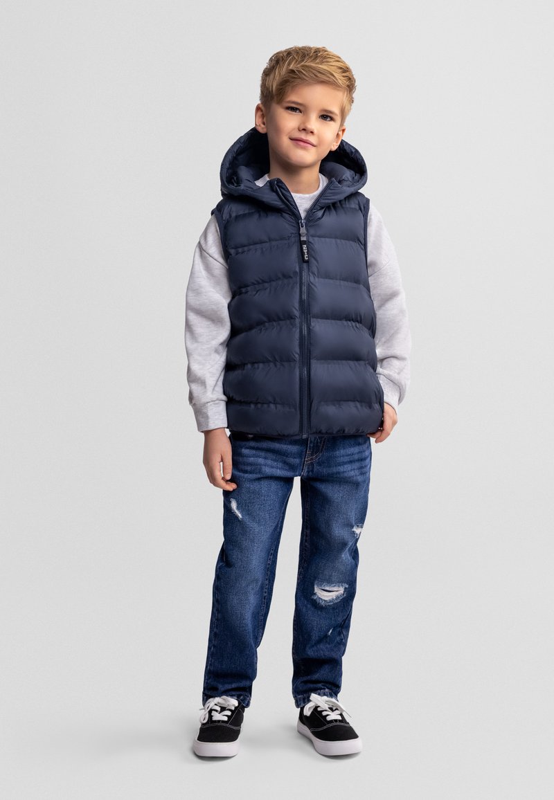 Jeune garçon debout portant un gilet matelassé bleu marine avec capuche, un sweat-shirt gris clair, un jean bleu déchiré et des baskets noires à lacets blancs.