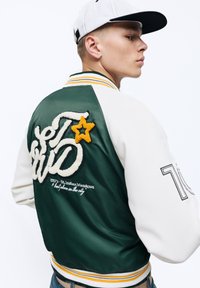 Groen en witte varsityjack met gele en witte borduursels, gestreepte ribgebreide manchetten en een opvallende sterdetail op de achterkant.