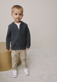 Pull gris à fermeture éclair avec une finition texturée, associé à un pantalon beige clair et des baskets blanches avec des accents rouges, debout à côté d'une table cylindrique dorée.