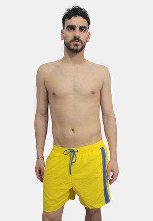 Pantaloni da bagno gialli realizzati in tessuto leggero, con coulisse blu e una striscia laterale con dettagli del logo. Design semplice, forma casual.
