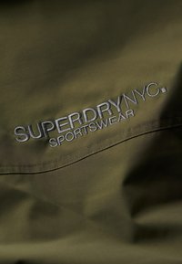 Olivenfarbener Stoff mit besticktem "SUPERDRY NYC. SPORTSWEAR"-Text in Weiß. Glatte Textur mit einem subtilen Glanz.