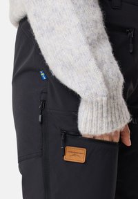 Svarta utomhusbyxor har en ficka med dragkedja och en läderetikett med "SWEDMOUNT". En ljus, stickad tröja syns från ärmslutet.