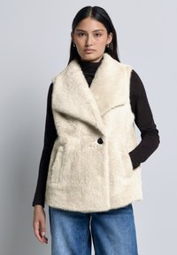 Gilet in finta pelliccia, di colore avorio, design senza maniche con colletto a scialle. Presenta una chiusura con un bottone e due tasche frontali. Texture morbida.