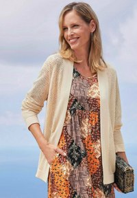 Beige-Mesh-Cardigan über einem mehrfarbigen, gemusterten Kleid mit floralen und geometrischen Designs, gehalten mit einer schwarzen, strukturierten Clutch.