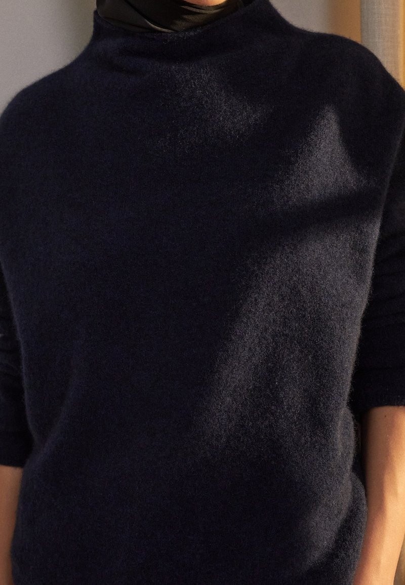 Personne portant un pull duveteux bleu marine foncé avec un col montant et des manches retroussées, baignée dans une lumière naturelle douce projetant des ombres.