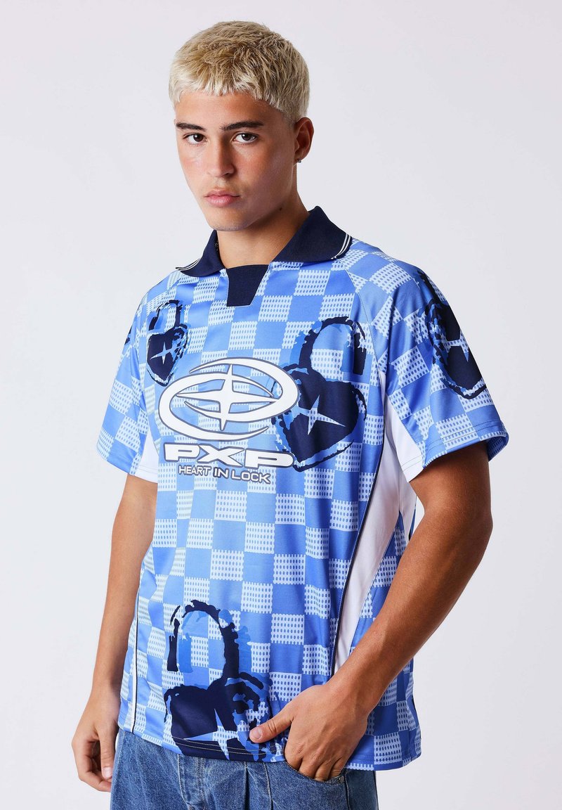 Jeune homme aux cheveux blonds courts portant une chemise à carreaux bleue avec des motifs de cadenas en forme de cœur foncé et le logo "PXP Heart in Lock".