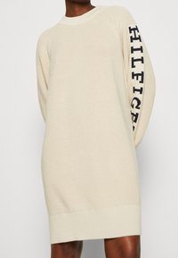 Abito maglione color crema con un motivo a trama e orlo a coste. Presenta un logo nero "HILFIGER" sulla manica sinistra.