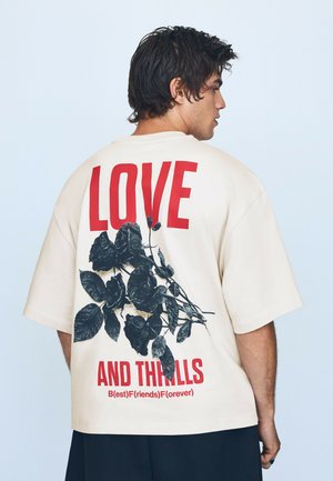 Mann mit dunklem Haar, der ein beige oversized T-Shirt mit rotem Text "LOVE AND THRILLS" und einer schwarzen Rosen-Grafik auf dem Rücken trägt.