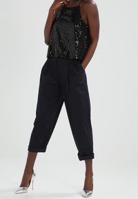 Crop top noir à sequins avec fines bretelles associé à un pantalon noir ample. Le mannequin porte des escarpins métalliques argentés à bout pointu.