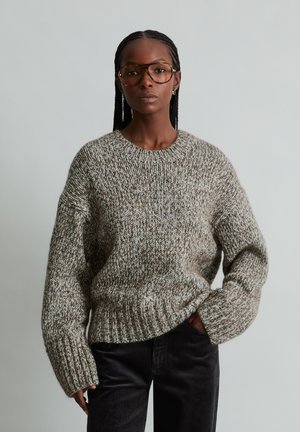 Frau mit geflochtenem Haar, die große Brillen, einen dicken grauen Strickpullover und schwarze Hosen vor einem einfachen hellen Hintergrund trägt.