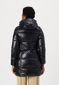 Zwarte pufferjas met een glanzende, gewatteerde buitenkant, capuchon en lange mouwen. Heeft een aansluitende taille en een knielange pasvorm.