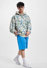 Hanorac cu glugă, cu un fundal de culoare deschisă, decorat cu motive florale albastre, asortat cu pantaloni scurți albaștri și adidași albi.