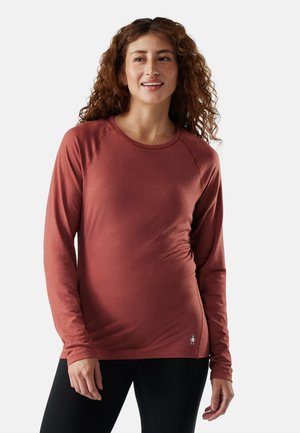 Femme aux cheveux bouclés portant un haut à manches longues couleur rouille et un pantalon noir, debout devant un fond clair uni.