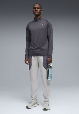 Man staat en draagt een grijze Puma top met lange mouwen, tweekleurige lichtgrijze broek, witte sneakers, en houdt een doorzichtige Puma waterfles vast.