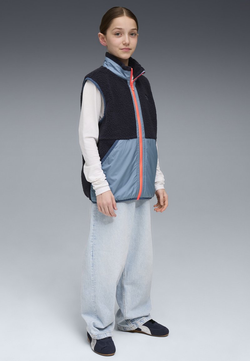 Navy- und blaue ärmellose Weste mit Fleece-Oberteil und kontrastierendem blauen Unterteil, ausgestattet mit einem orangefarbenen Reißverschluss. Weite hellblaue Jeans.