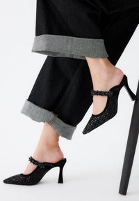 Mules negros abiertos en la punta, con una puntera puntiaguda, un diseño tejido texturizado y embellishments, combinados con pantalones de mezclilla oscuros arremangados en los puños.