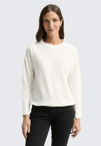 Pull blanc avec un design en tricot texturé, col rond et manches longues, associé à un pantalon noir. Silhouette simple et moderne.