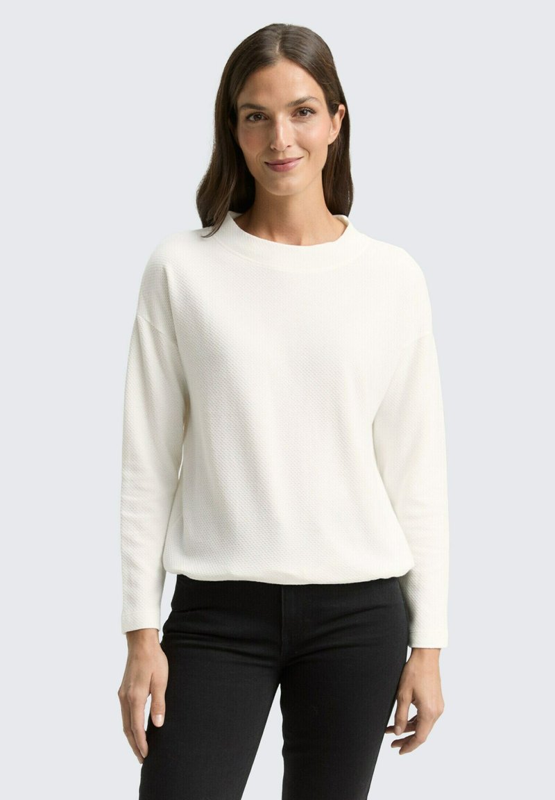 Pull blanc avec un design en tricot texturé, col rond et manches longues, associé à un pantalon noir. Silhouette simple et moderne.