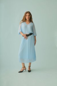 Femme debout devant un fond bleu pâle, portant une robe midi plissée bleu clair avec des manches transparentes, une large ceinture noire et des escarpins noirs à bride cheville.