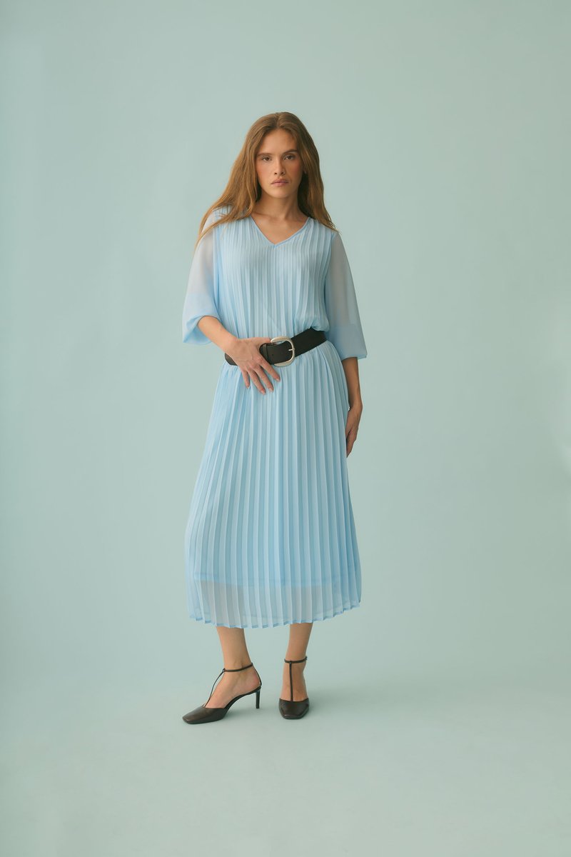 Femme debout devant un fond bleu pâle, portant une robe midi plissée bleu clair avec des manches transparentes, une large ceinture noire et des escarpins noirs à bride cheville.