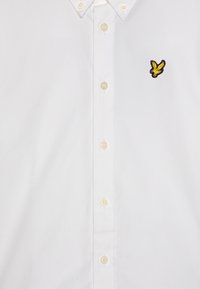 Hvit skjorte med spredt krage, med en gul og svart brodert fuglelogo på venstre bryst og beige knapper.