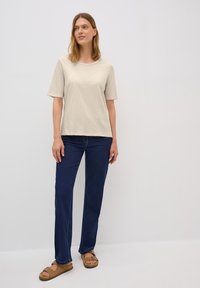 Beige tričko s krátkým rukávem, volný střih, v kombinaci s tmavě modrými džínami rovného střihu a hnědými sandály se stahovací šněrovací designem na prstech.
