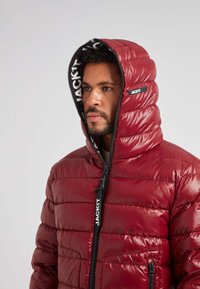 Rød puffjakke med en skinnende finish, vandret syninger, høj krave og sort lynlås med branding. Hætten er delvist lynet op.