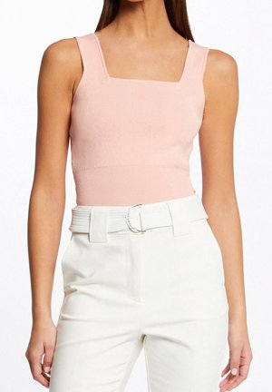 Frau trägt ein ärmelloses, quadratisches Ausschnitt rosa Stricktop kombiniert mit hochtaillierten weißen Hosen und einem passenden weißen Gürtel mit silberner Schnalle.