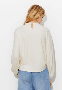 Blouse crème à manches longues avec une surface texturée, un col à volants et un petit détail en forme de trou de serrure au dos. Associée à un jean bleu clair.