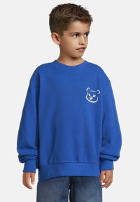 Blå bomullströja med lång ärm, ribbad mudd och fåll. Utrustad med ett vitt björngesicht broderat på vänster bröst. Avslappnad passform.