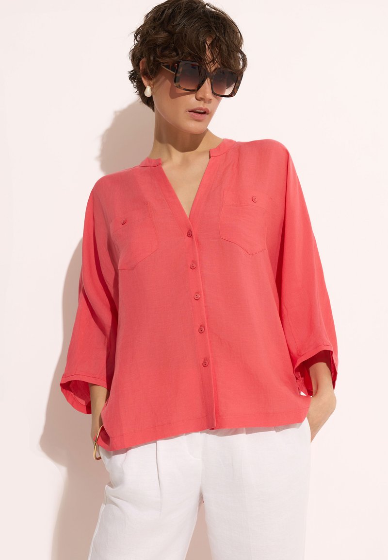 Koronkowa bluzka z guzikami w kolorze koralowym, z dekoltem w serek, podwiniętymi rękawami i dwiema kieszeniami na piersi, wykonana z lekkiego materiału. Stylizowana z białymi spodniami.