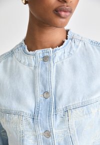 Ljusblå denimjacka med fransigt krage, silverknappar och broderade detaljer på framsidan av fickan.