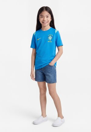 Enfant souriant debout, une main dans la poche, portant un maillot de football du Brésil bleu, un short en jean et des chaussures blanches sans lacets sur fond blanc.
