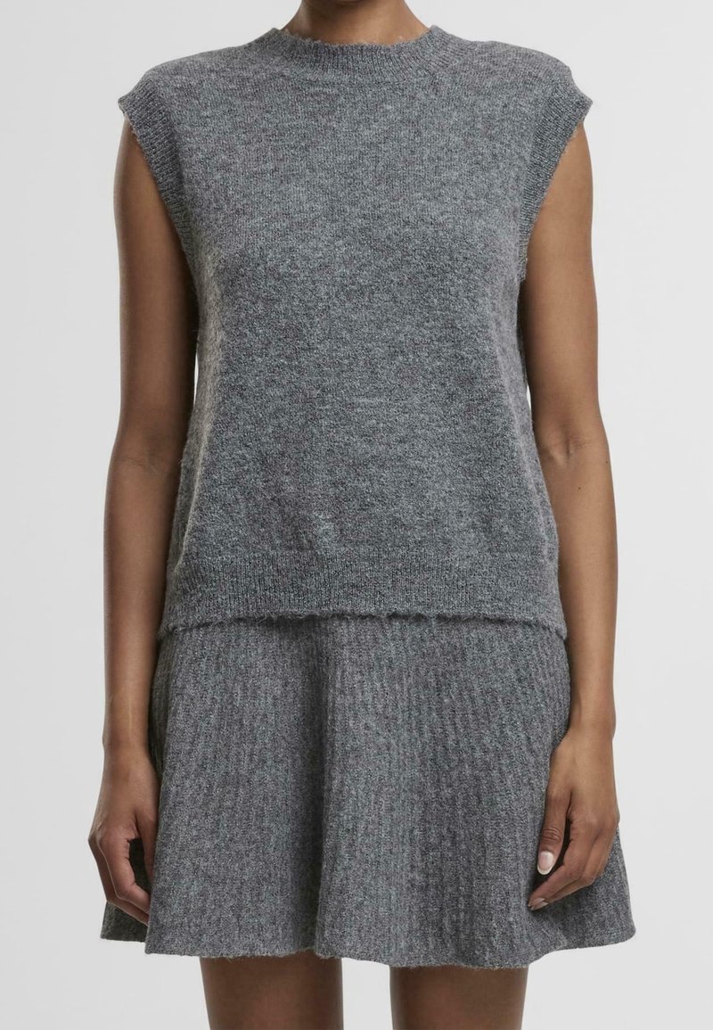 Femme portant un haut en tricot gris sans manches et une jupe en tricot gris assortie sur un fond uni.