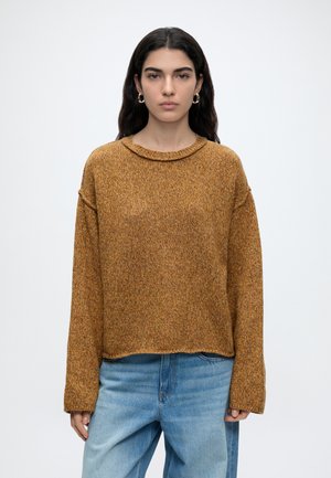 Femme aux longs cheveux foncés portant des boucles d'oreilles créoles dorées, un pull en maille marron ample et un jean bleu clair, debout devant un fond blanc uni.