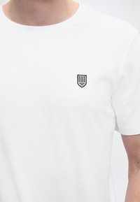 Bijela pamučna majica kratkih rukava s malim crnim vezanim logotipom štita na lijevoj strani prsa, okruglim izrezom na vratu i kratkim rukavima.