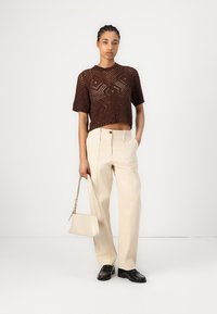 Maglione cropped marrone a maglia con maniche corte, pantaloni beige a vita alta con una vestibilità rilassata, scarpe nere con lacci e una borsa leggera.