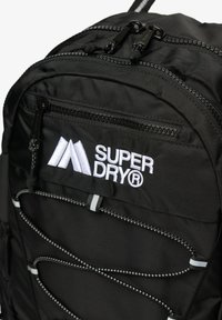 Schwarzer Rucksack mit einem auffälligen weißen Logo, Reißverschlusstaschen, elastischen Bungee-Kordeln und strukturiertem Stoff.