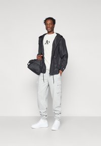 Casaco leve preto com capuz, t-shirt creme com texto, calças de moletom cinzas com bolsos e ténis brancos, segurando uma bolsa preta.