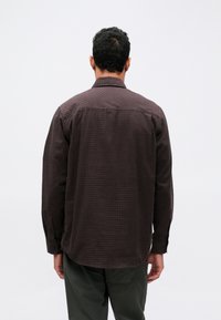 Tumepruun ruuduline pika varrukaga särk klassikalise kraega, nuppudega mansetid ja vabalt langev lõige, koos oliivroheliste pükstega.