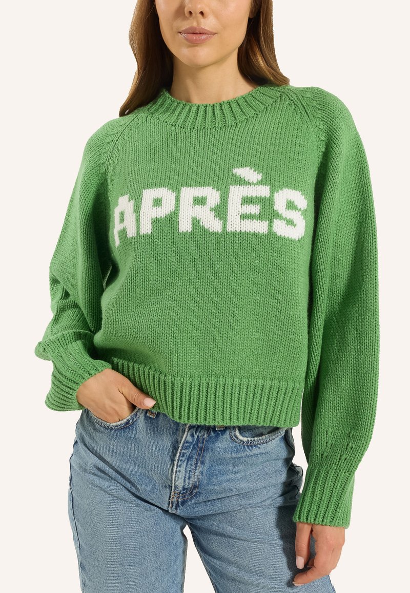 Grüner Strickpullover mit weißem "APRÈS"-Text. Gerippter Rundhalsausschnitt und Bündchen. Kurz geschnittenes Design mit lockerer Passform, kombiniert mit blauen Jeans.