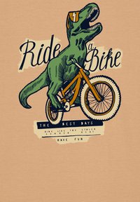 Grafisches T-Shirt mit einem grünen Dinosaurier, der ein oranges Fahrrad fährt. Der Text lautet "Fahr ein Fahrrad" und weitere Phrasen in Schwarz. Beiger Hintergrund.