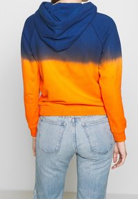 Osoba v modro-oranžovém ombre mikině s kapucí dolů a světlých modrých džínách, zobrazená zezadu.