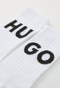 Egy pár fehér bordás zokni, nagy fekete betűkkel írva "HUGO", sík felületen elhelyezve.