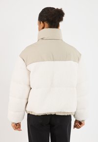 Ocieplana kurtka z beżową górną częścią i białym teksturowanym materiałem na dole. Posiada wysoki kołnierz i elastyczne mankiety, a także krótki krój.