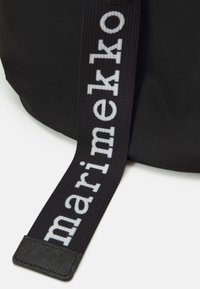 Black bag with a textured surface, featuring a wide strap displaying "marimekko" in bold white letters. Strap has a leather accent.