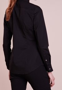 Person klädd i en åtsittande svart långärmad skjorta och svarta byxor, sedd bakifrån mot en ljusbeige bakgrund.