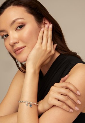 Mujer con cabello largo y castaño que lleva un top negro sin mangas, apoyando su rostro en una mano y mostrando una pulsera de plata con dijes.