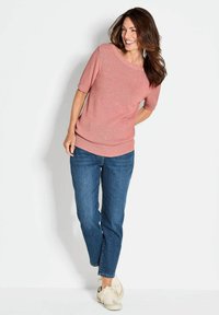 Lichtroze korte mouw trui met een bootnek, gecombineerd met middendonkere blauwe rechtpassende jeans en witte sneakers, staand tegen een effen achtergrond.